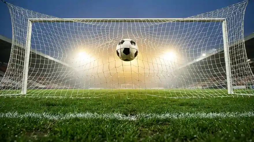 Pallone che entra nella rete di una porta di calcio su campo in erba durante una partita serale allo stadio
